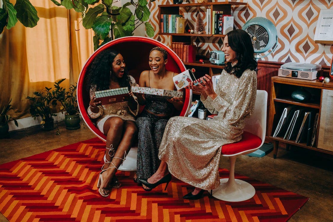 Three women in festive outfits open smart home gifts in a modern-retro-style, orange-and-red den.
