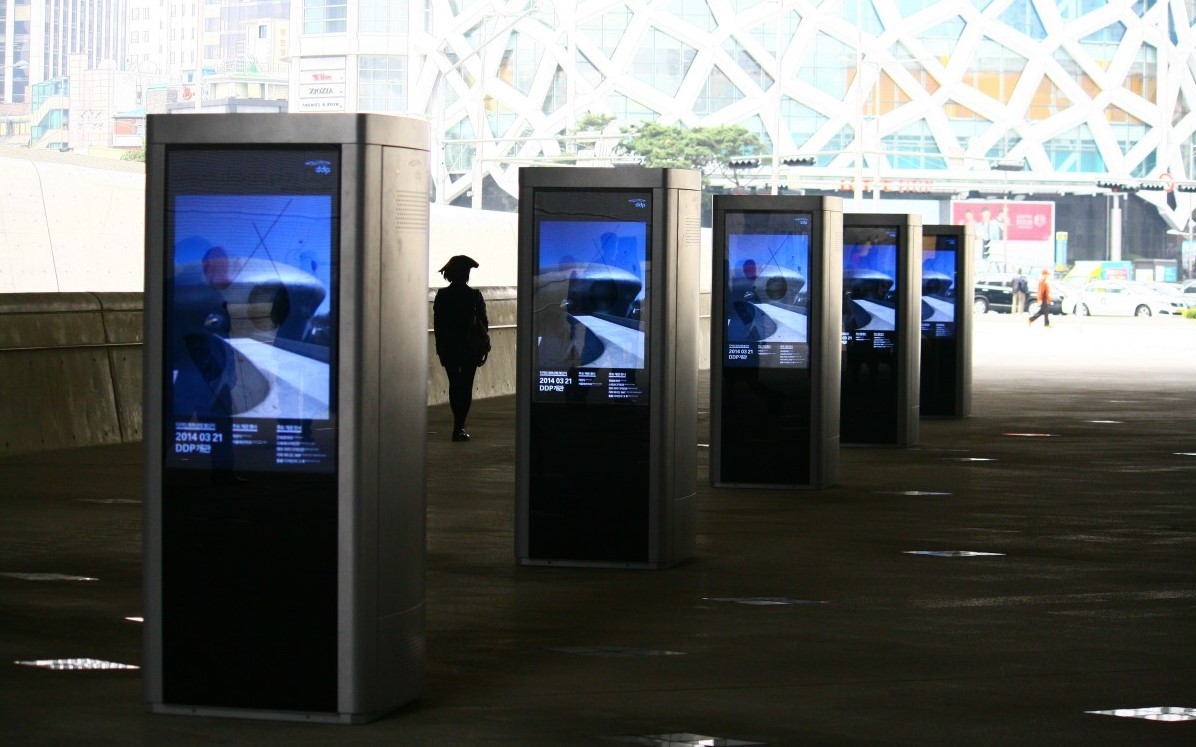 Digital wayfinding signage kiosks greet guests at a sunny outdoor plaza, with shops in the background