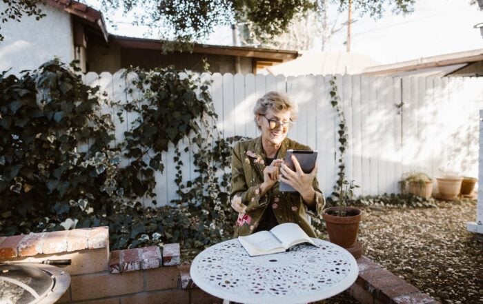 An older woman sits on her bright patio and uses a tablet to check how smart home energy solutions have saved her money.