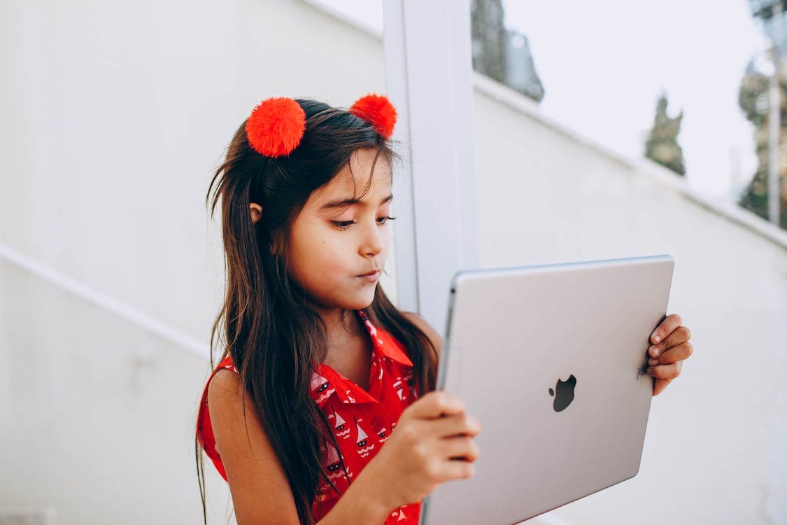 A young girl wearing a bright red-orange shirt and hair accessories watches a sleek tablet using home automation.