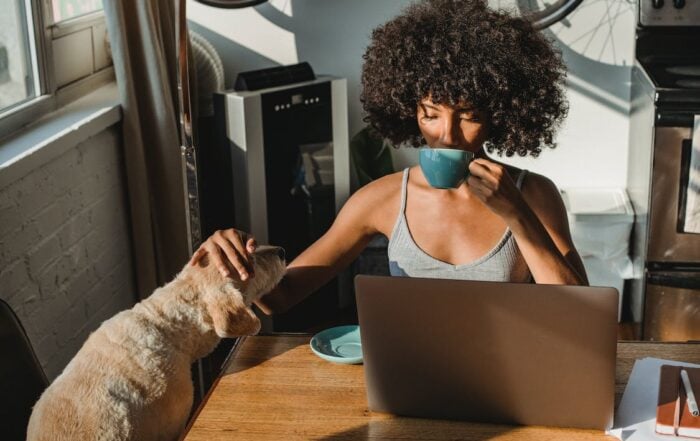 A woman uses smart home voice control features in front of a laptop while drinking coffee and petting a small dog. The room is full of sunshine and neutral colors.