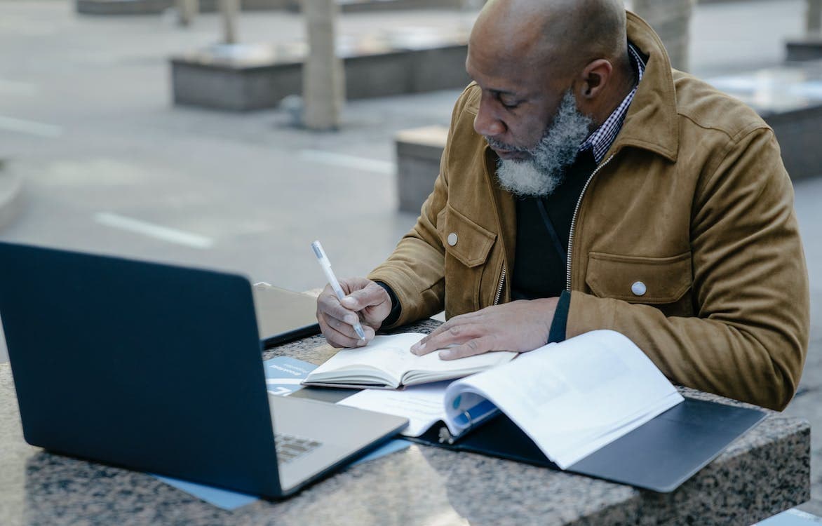A man sits at a table outdoors with a laptop in front of him